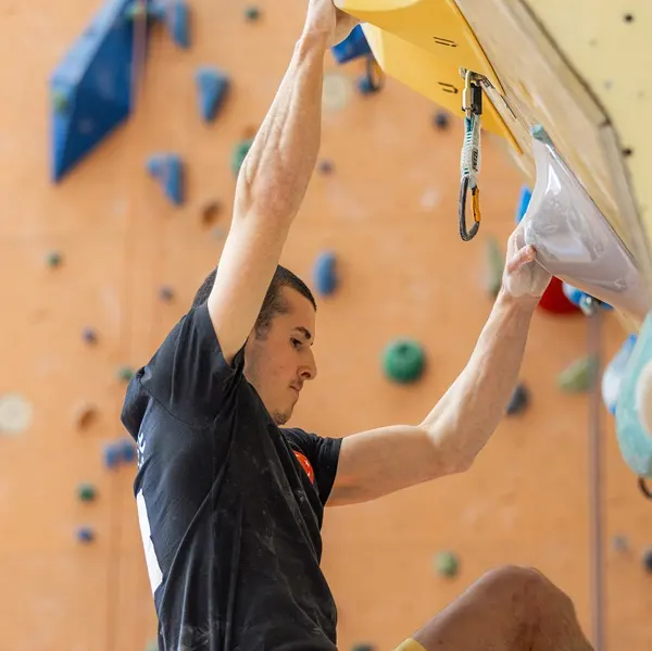 Een man in een zwart shirt beklimt een indoor-klimwand en houdt zich met beide handen vast aan een grote greep. Op de achtergrond zijn verschillende gekleurde grepen te zien op een oranje wand.